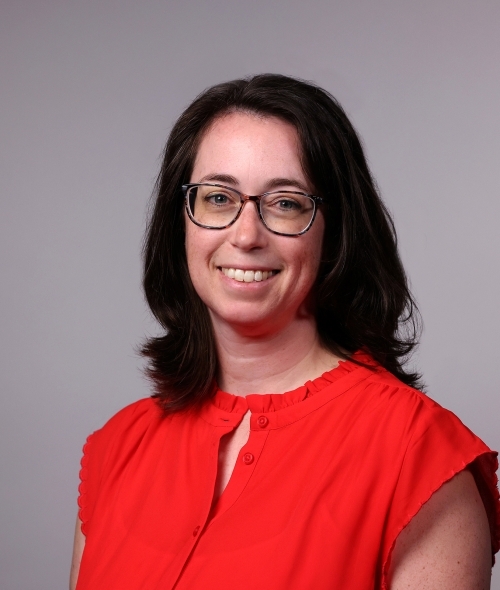 a woman with curly brown hair wearing glasses and a red top on a grey background