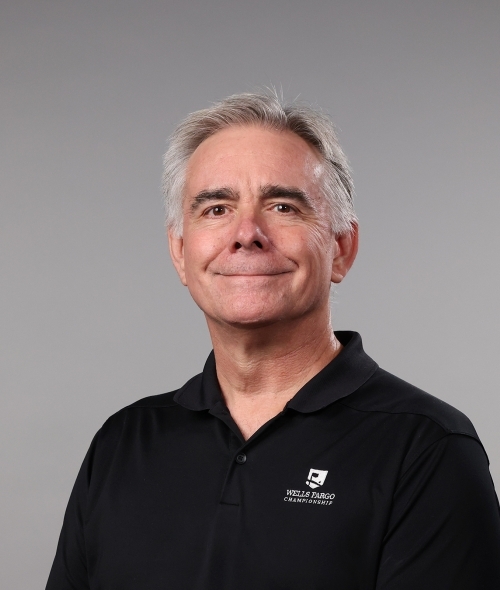 a man with short white hair wearing a collared black shirt and smiling on a grey background
