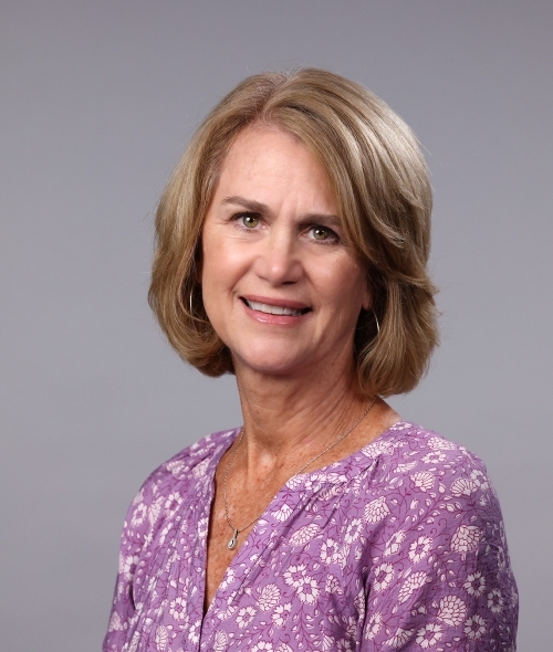 a woman with short blonde hair wearing a floral purple top on a grey background