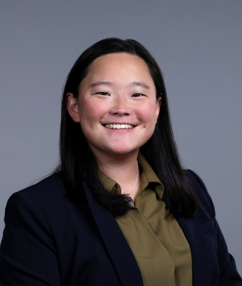 Smiling headshot of seated woman with black hair, green button down shirt, black blazer
