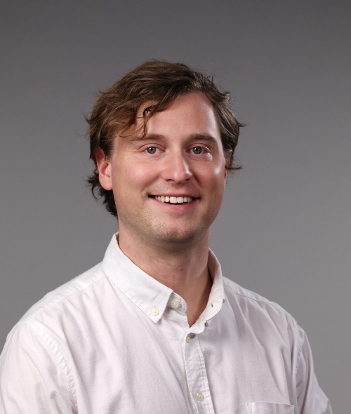 a professor with short dirty blonde hair wearing a white collared shirt smiling on a grey background