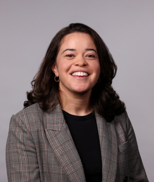 a professor with curly dark hair wearing a black top and grey blazer on a grey background