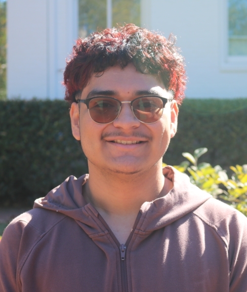 a young man with reddish brown hair wearing glasses and a sweatshirt standing outside on a sunny day
