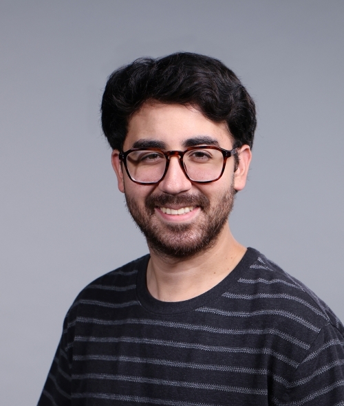 a man with dark hair wearing glasses and a black striped shirt on a grey background