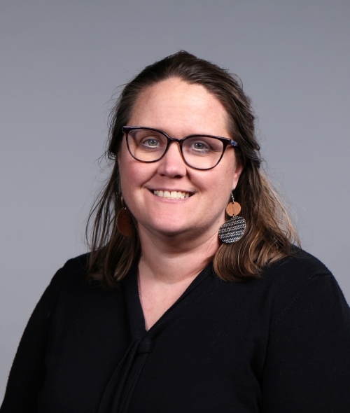 a woman with curly dark hair wearing glasses and a black blouse with large earrings on a grey background