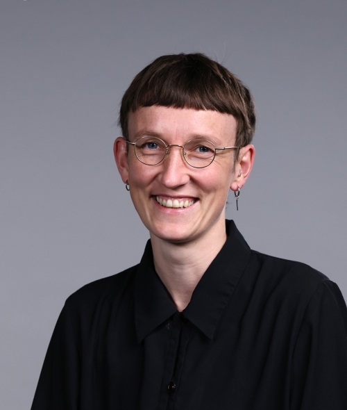 a young person with short hair wearing glasses and a black blouse on a grey background