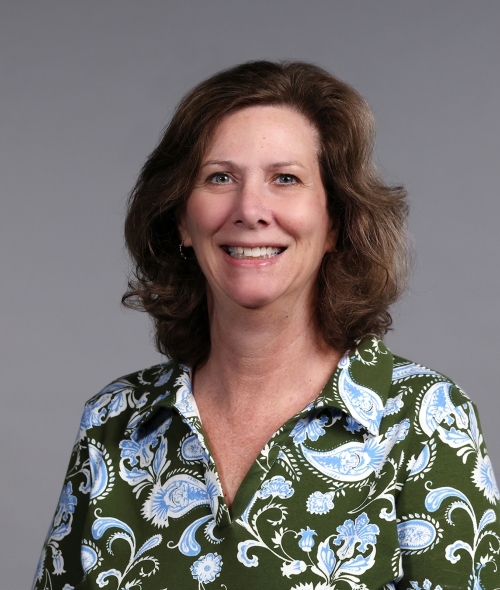 a woman with curly brown hair wearing a floral green and blue blouse on a grey background