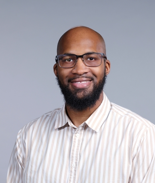 a Black man with glasses and facial hair wearing a collared white shirt on a grey background