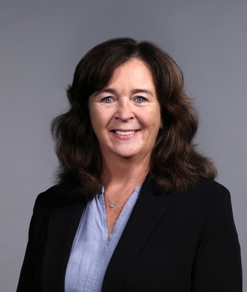 a woman with curly dark hair wearing a blue blouse and black blazer on a grey background