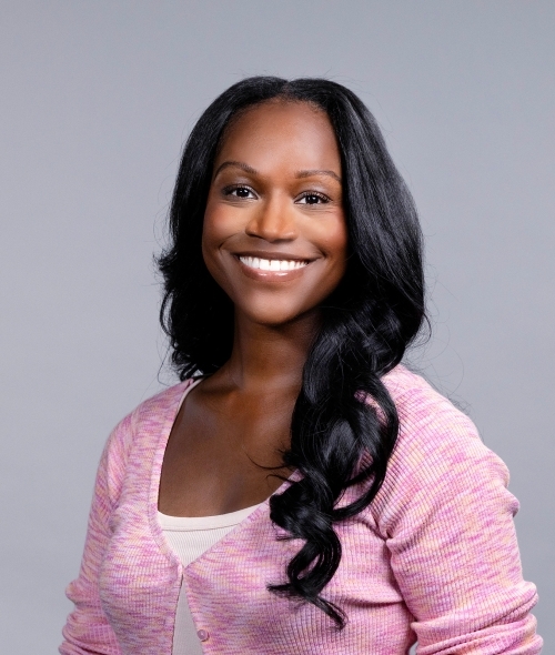 a young woman with curly black hair wearing a pink cardigan on a grey background