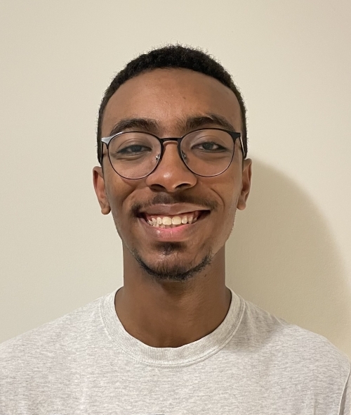 Young adult man smiles with short black hair, medium skin tone, and black facial hair and glasses.