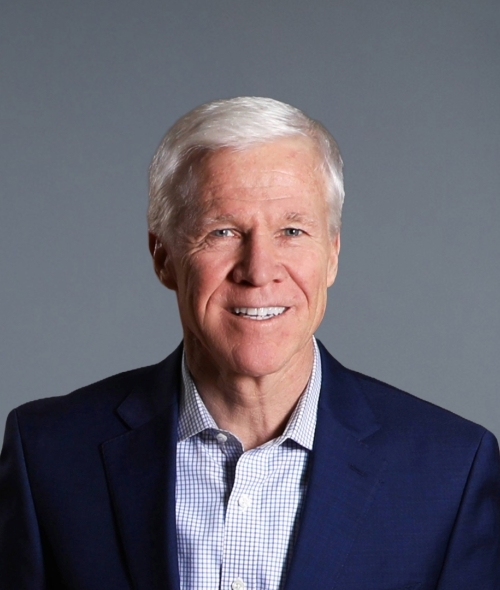 a white man with white hair wearing a collared shirt and jacket on a grey background