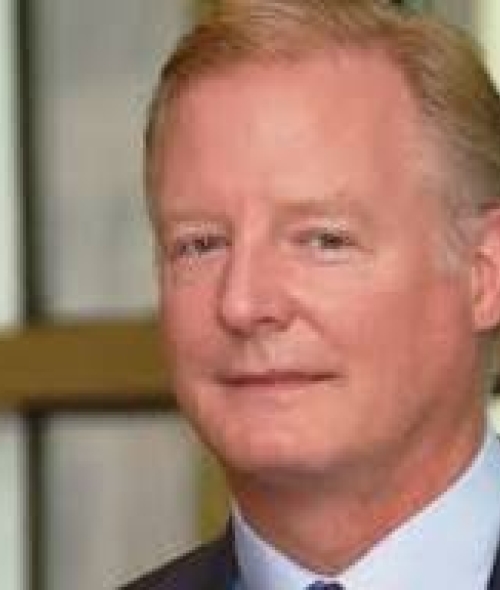A man with light hair, wearing a dark suit jacket and a light blue collared shirt, looks forward with a neutral expression.