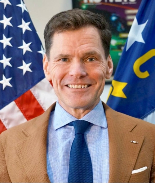 Grier Martin smiles while wearing a tan blazer, light blue patterned shirt, and dark blue tie, positioned between the United States flag and the North Carolina state flag.