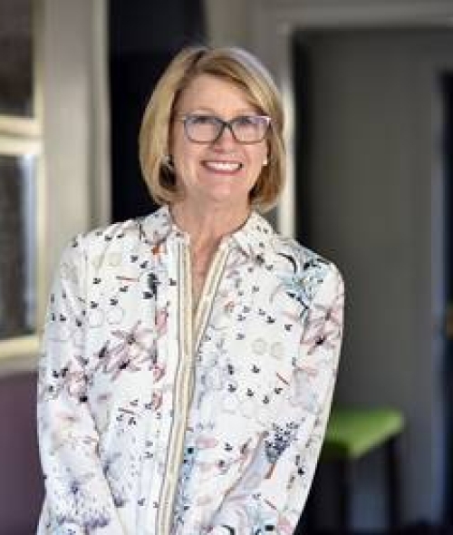 Katie Belk-Morris smiles while wearing glasses and a white button-down shirt with a floral and bird pattern.