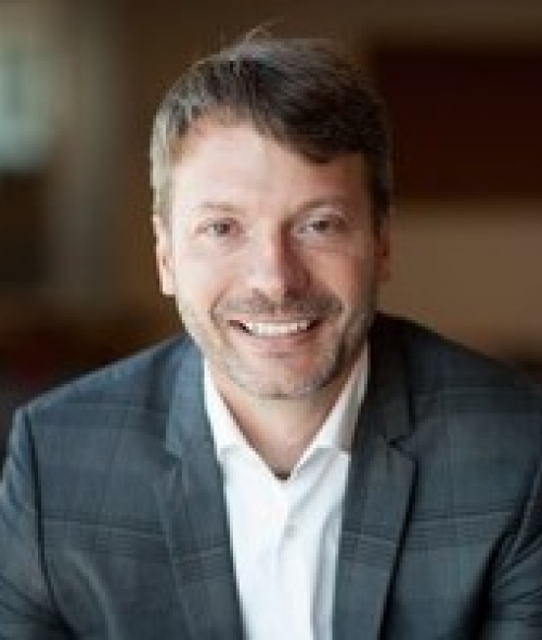a smiling man with short brown hair and a light beard, wearing a grey plaid blazer over a white shirt.