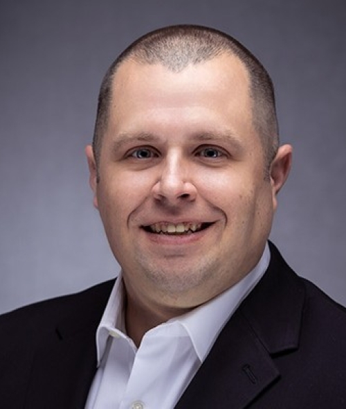 A headshot of a smiling man with short-cropped hair, wearing a white button-down shirt and a black blazer against a neutral grey background.