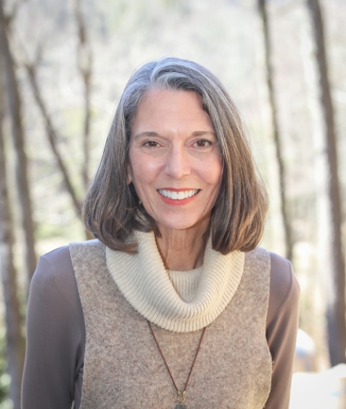 A smiling woman with shoulder-length gray hair wearing a tan cowl-neck sweater and a stone pendant necklace, standing outdoors with a blurred forest background.