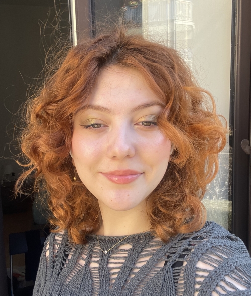 A woman with wavy, vibrant ginger hair and a soft smile looks directly at the camera while wearing a grey crocheted top and shimmering gold eyeshadow.