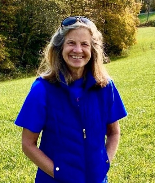 A smiling woman with light brown hair and sunglasses on her head stands in a sunlit green field wearing a bright blue short-sleeved puffer vest.