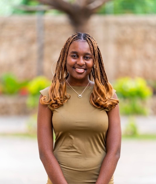 a young Black woman wearing a green dress and standing outside in front of a brick wall with flowers in the distance