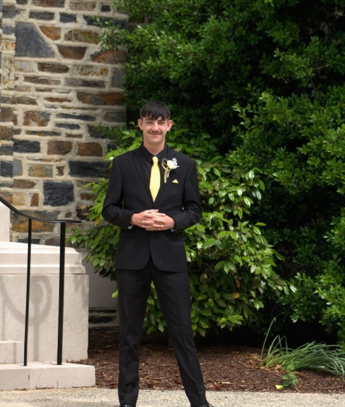 a young man wearing a black suit and yellow tie standing in front of a brick building with green bushes around