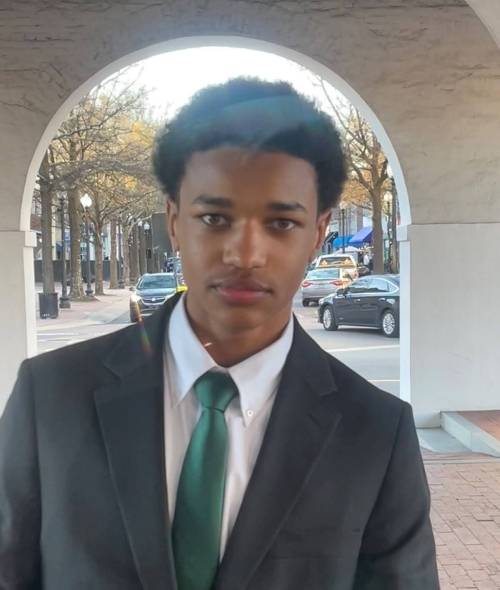 a young Black man wearing a suit and tie standing in front of a large stone arch in front of a busy city street
