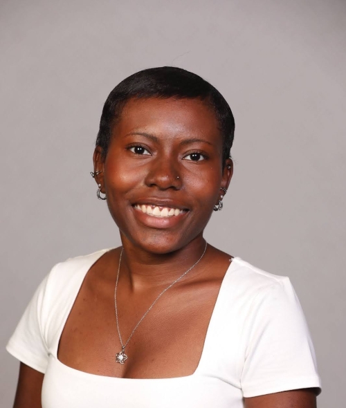 A smiling Black woman with short hair, wearing a white square-neck top, a silver flower pendant necklace, and hoop earrings, posing against a neutral gray background.