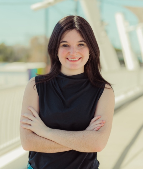 A smiling young woman with dark shoulder-length hair and freckles stands with her arms crossed on a sunlit pedestrian bridge with white railings and modern architectural details.