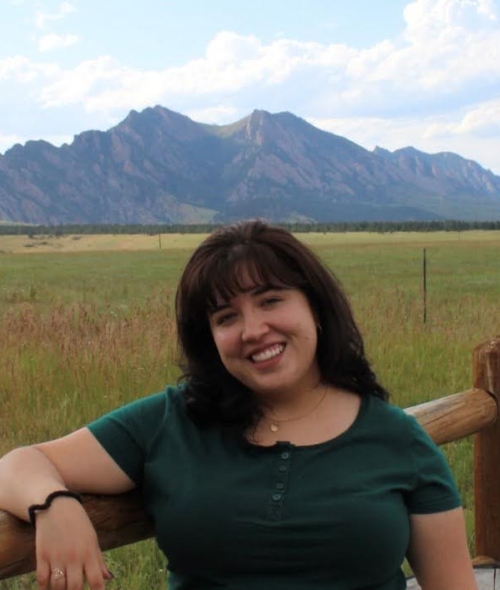 Caroline Evans smiles while leaning on a wooden fence with a vast green field and a jagged mountain range under a cloudy sky in the background.