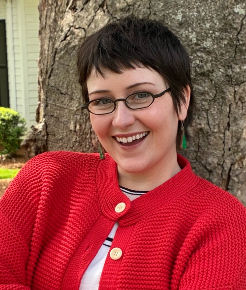 A young woman with a dark brown pixie cut, black round glasses, and a red sweater smiles outdoors