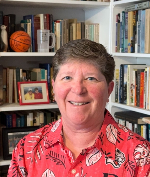 A person with short, brown hair and a smiling expression wears a red patterned collared shirt while posing in front of white bookshelves filled with books, a small basketball, and a framed photograph.