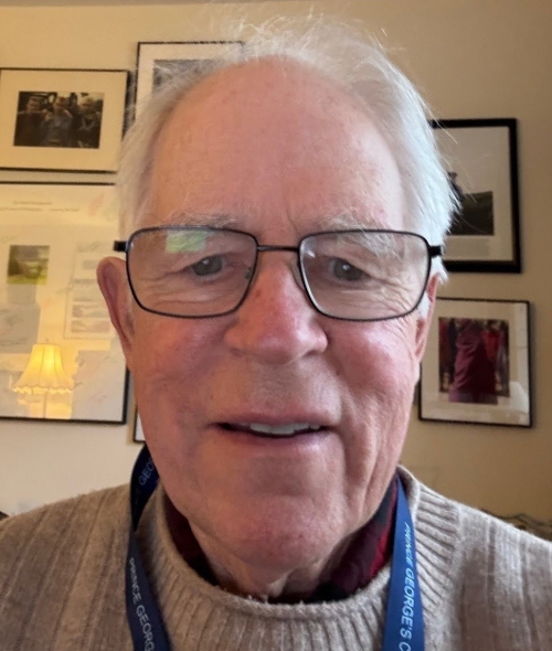 A close-up headshot of Joe Howell wearing glasses, a tan sweater, and a blue lanyard, with framed pictures on the wall behind him.