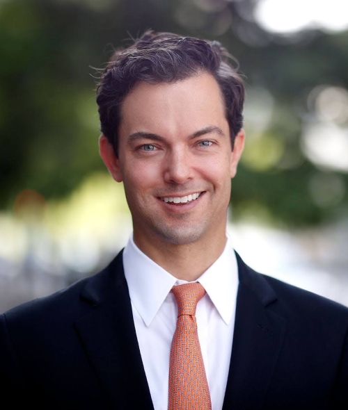 A professional portrait of P.J. Martin wearing a black suit, white shirt, and orange patterned tie against a softly blurred outdoor background.