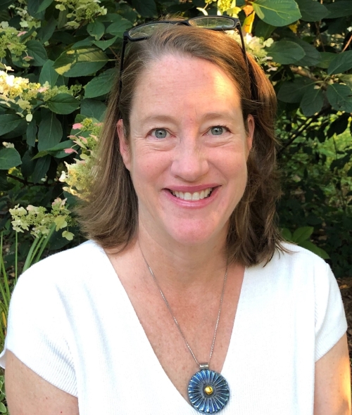 A smiling adult white woman with shoulder-length reddish brown hair, a white shirt, and a large pendant necklace in front of a lush green background
