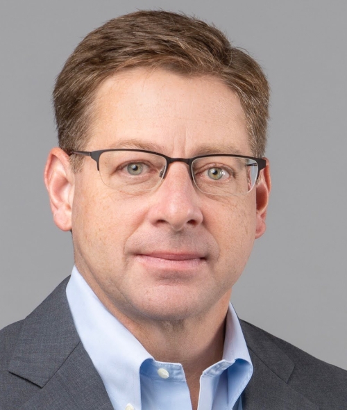 A headshot of Winn Maddrey wearing glasses, a light blue dress shirt, and a grey suit jacket against a solid grey background.