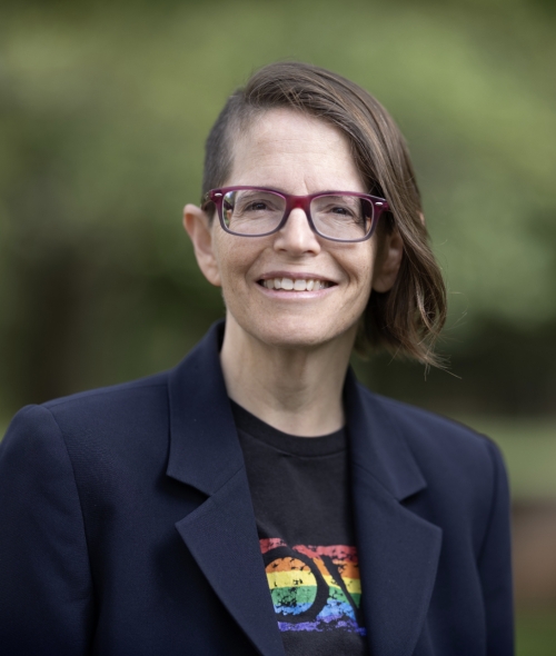 A person with brown, wavy hair in an asymmetrical style wears a blue blazer, glasses, and graphic tee
