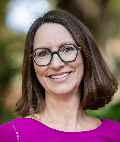 A woman with shoulder-length brown hair and glasses and a bright purple shirt