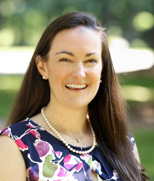 A smiling woman with long brown hair, pearl earrings, and a pearl necklace, wearing a multi-colored floral sleeveless top against a blurred green outdoor background.