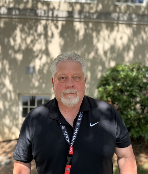 man with grey hair and a goatee wearing a black Nike polo shirt and a "Wildcats" lanyard, pictured from the waist up outdoors in front of a building.