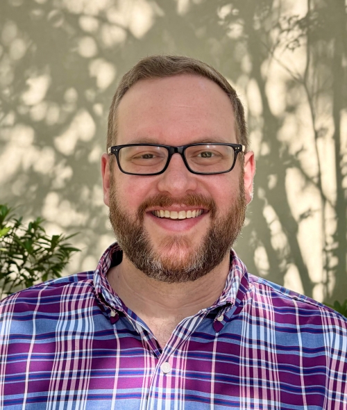 smiling man with glasses and a beard wearing a purple and blue plaid button-down shirt, pictured from the chest up against a sunlit wall with leafy shadows.