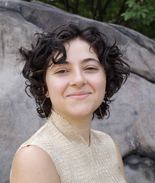 A person with short dark curly hair and a tan blouse