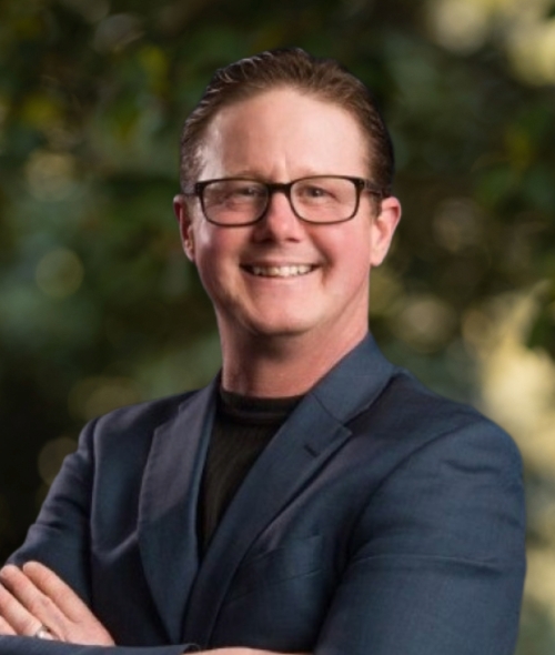 A professional headshot of a smiling man with glasses, wearing a dark blue blazer over a black shirt, posing with his arms crossed against a blurred green background.