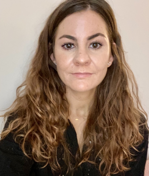 A woman with long, wavy brown hair facing forward with a neutral expression against a plain background.