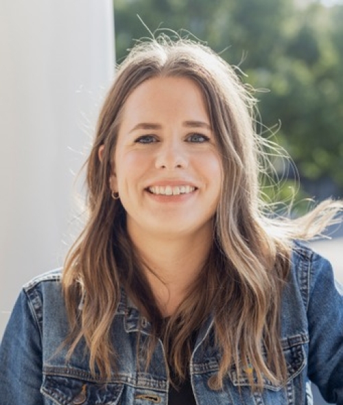 A smiling woman with light brown, shoulder-length hair and blue eyes wears a denim jacket over a black shirt while posing outdoors.