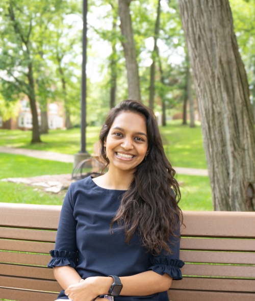 A smiling young woman with long, dark, wavy hair sits on a wooden park bench outdoors, wearing a dark blue blouse and white pants, with a green, tree-lined campus lawn blurred in the background.