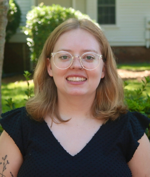 A headshot of a smiling woman with shoulder-length blonde hair, round clear-framed glasses, and a black textured top, posed outdoors against a soft-focus background of green foliage and a house.