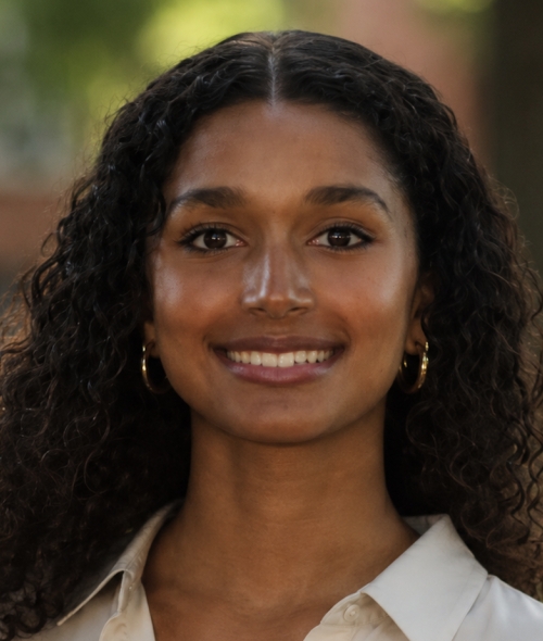 A smiling young woman with long, curly dark hair and gold hoop earrings, wearing a light-colored collared shirt against a softly blurred natural background.