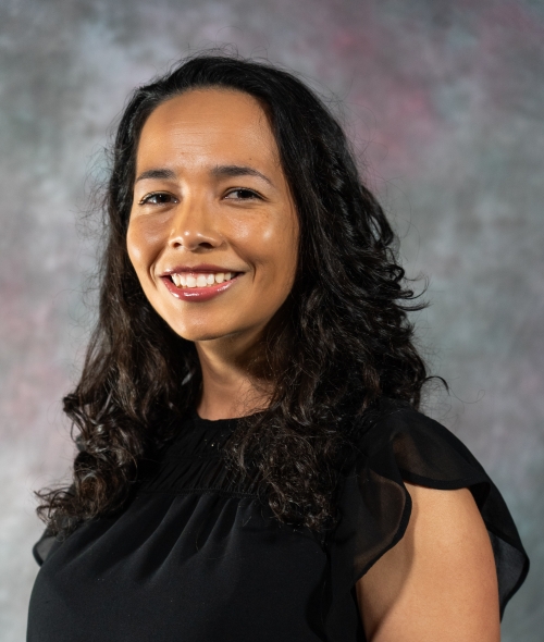 A professional headshot of a smiling woman with long, dark curly hair, wearing a black sleeveless blouse against a textured gray background.