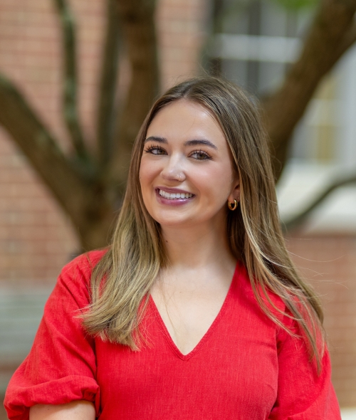 Amelia Solum smiling in a red V-neck top with a blurred brick building and tree in the background.